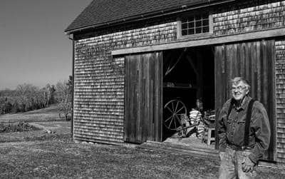 BILL KENDALL stands near the barn at Kendall farm, with his great-grandmother’s spinning wheel just inside the barn door. The rebuilding of the kitchen in the 200-year-old farmhouse is trigg