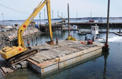LOADING AN EXCAVATOR on a barge to dig out the fill in the Eastport breakwater, a portion of which collapsed in December, is a crew from contractor CPM Constructors. Work on the $14.95 milli